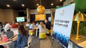 Group of educators seated in casual modern workspace with colorful ottomans during ECO Center AI Summit presentation, with speaker at front and branded event signage visible