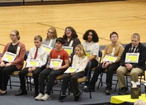 Alt Text: A group of students sit in chairs on a gymnasium floor wearing numbered "Participant" signs for a spelling bee competition. The group includes both younger and older students dressed in formal attire.