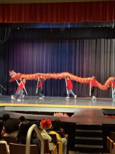 Students in red shirts perform a traditional Chinese dragon dance on a school auditorium stage, carrying a long red and gold dragon prop on poles before a seated audience.