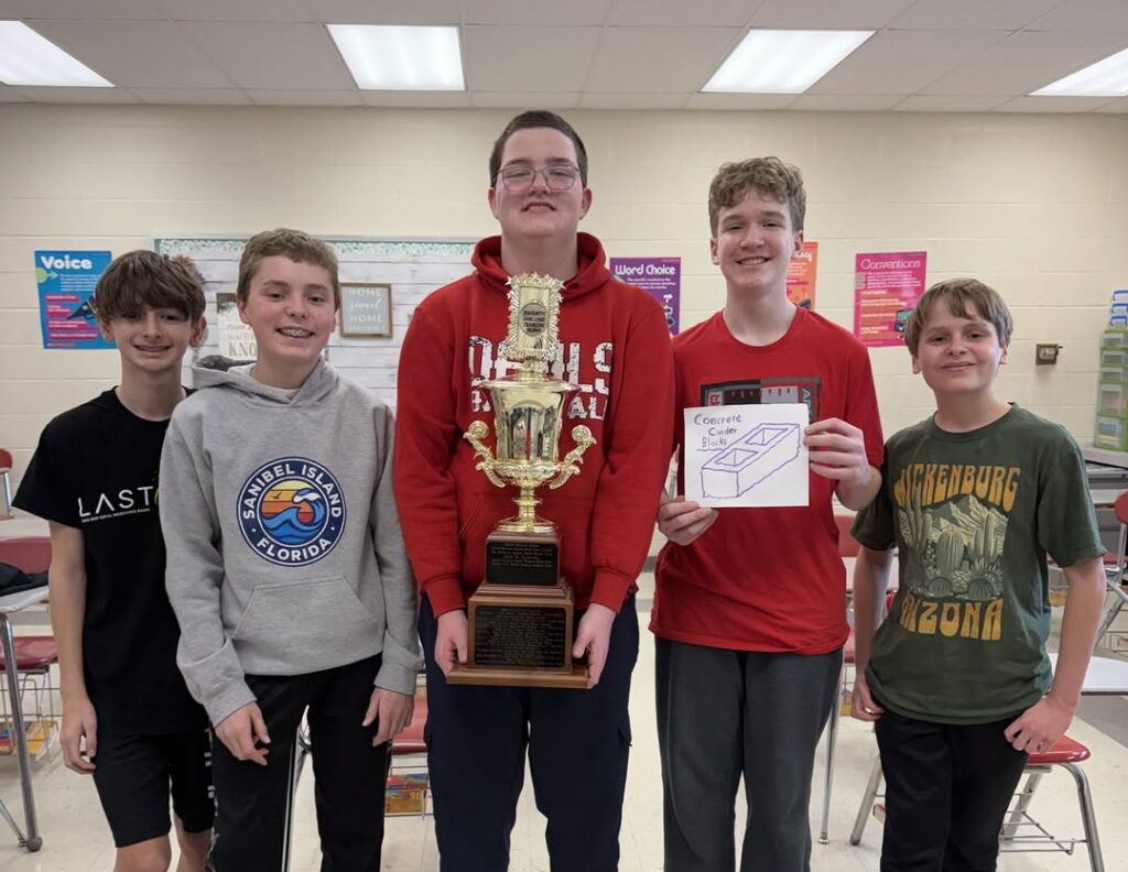 Five middle school students stand together in a classroom holding a large gold trophy and a hand-drawn sign reading "Concrete Cinder Blocks."