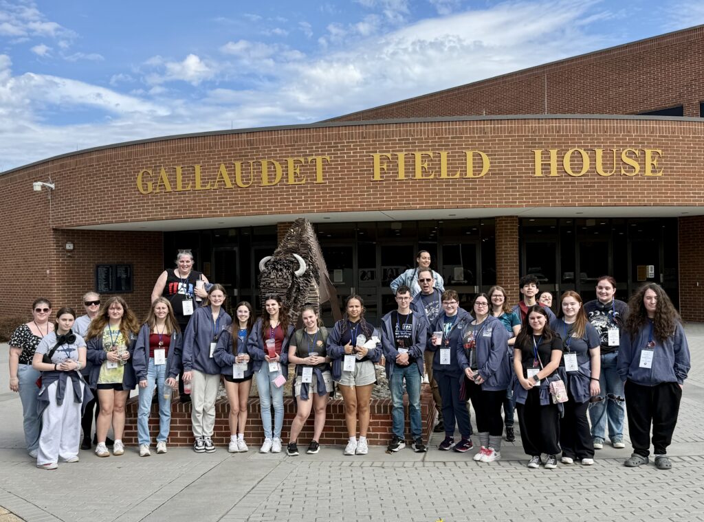 A large group of Ohio ASL students and chaperones pose for a group photo in front of the Gallaudet Field House, a brick building with gold lettering, on the campus of Gallaudet University in Washington, D.C.