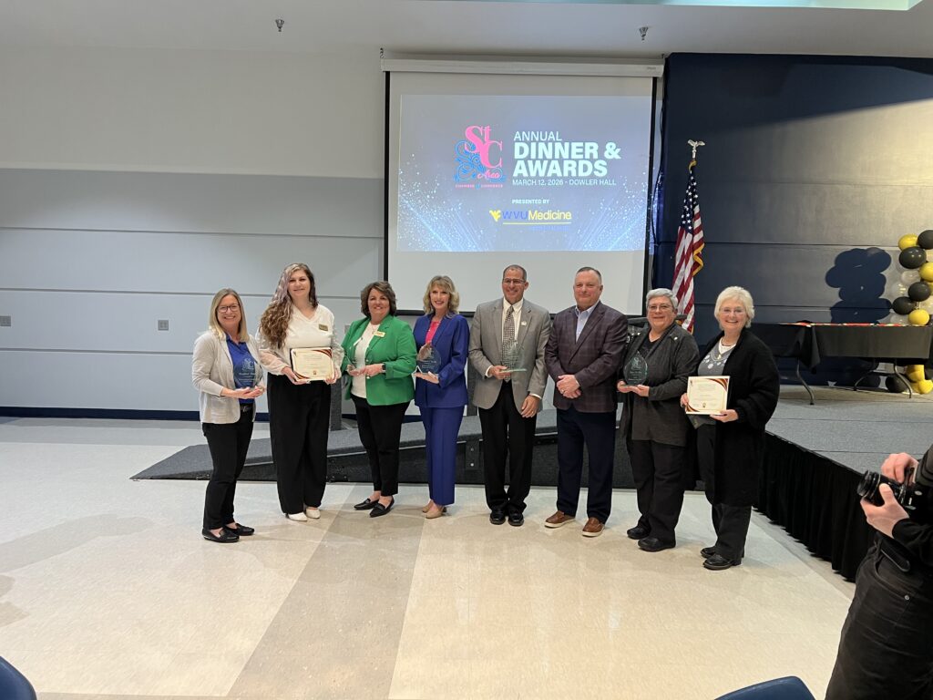 A group of eight award recipients pose for a photo at the St. Clairsville Area Chamber of Commerce 2026 Annual Dinner and Awards program at Dowler Hall. Several individuals hold glass trophies and certificate folders. A projection screen displaying the event name and date of March 12, 2026, is visible in the background.
