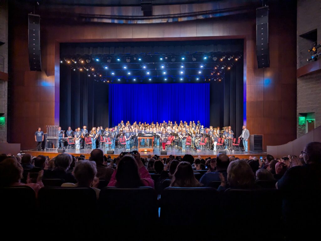 A large group of middle school students in matching dark T-shirts stands and sits on a performing arts center stage in front of a royal blue curtain backdrop, with musical instruments and red chairs visible. A full audience fills the seats in the foreground.