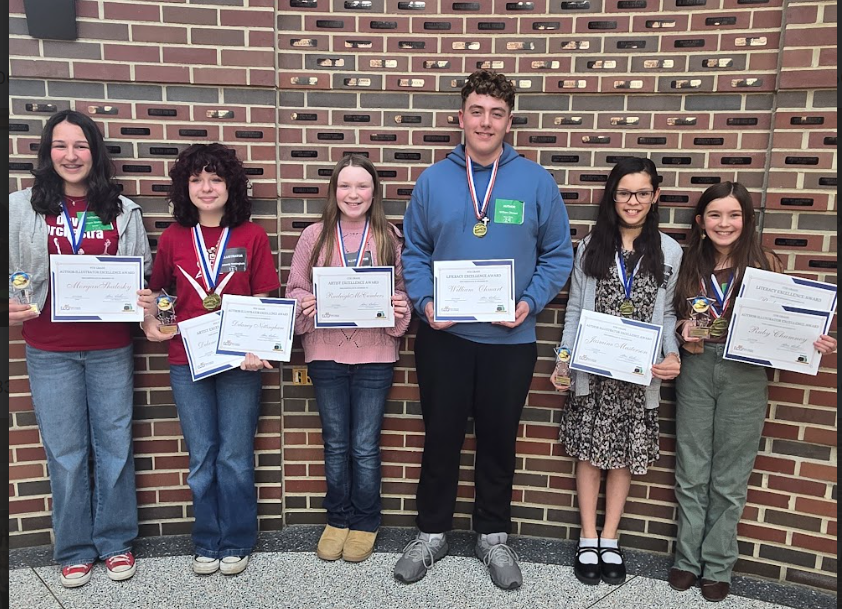 Seven students stand in a row in front of a brick wall, each holding certificates and medals from the Young Authors and Illustrators competition. Pictured from left to right: Morgan Shalosky, Delaney Nottingham, Raeleigh McCumbers, William Omhart, Jasmine Masterson, and Ruby Chumney.