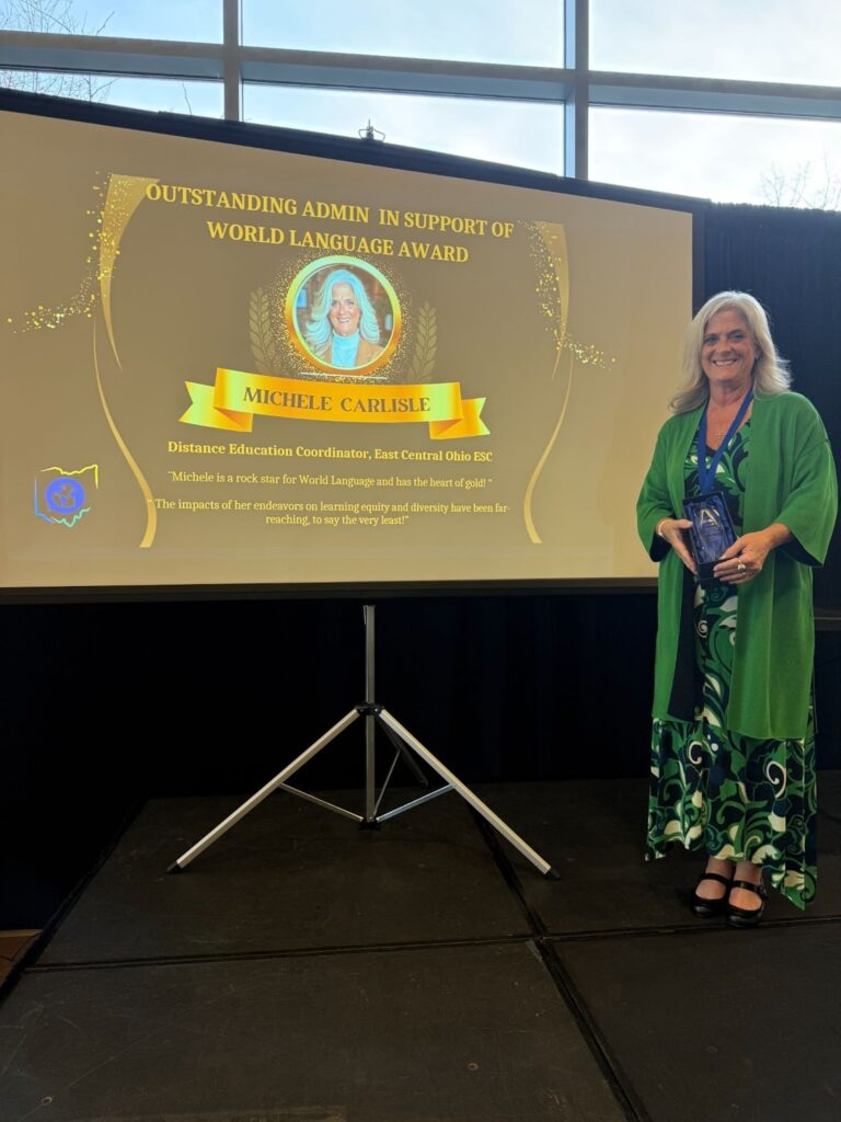 Michele Carlisle, Distance Education Coordinator at East Central Ohio ESC, stands smiling beside a presentation screen displaying her photo and the "Outstanding Admin in Support of World Language Award" from the Ohio World Language Association.