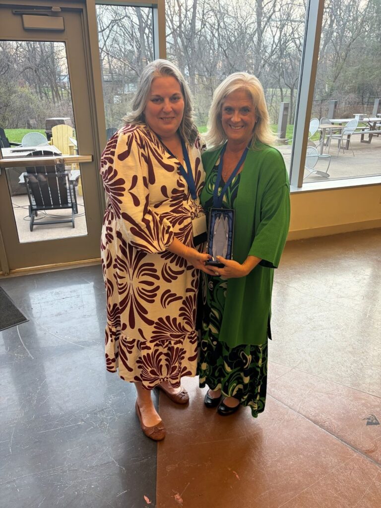 Two women stand together smiling and holding a crystal award trophy. The woman on the left wears a cream and burgundy floral dress; the woman on the right wears a green outfit.
