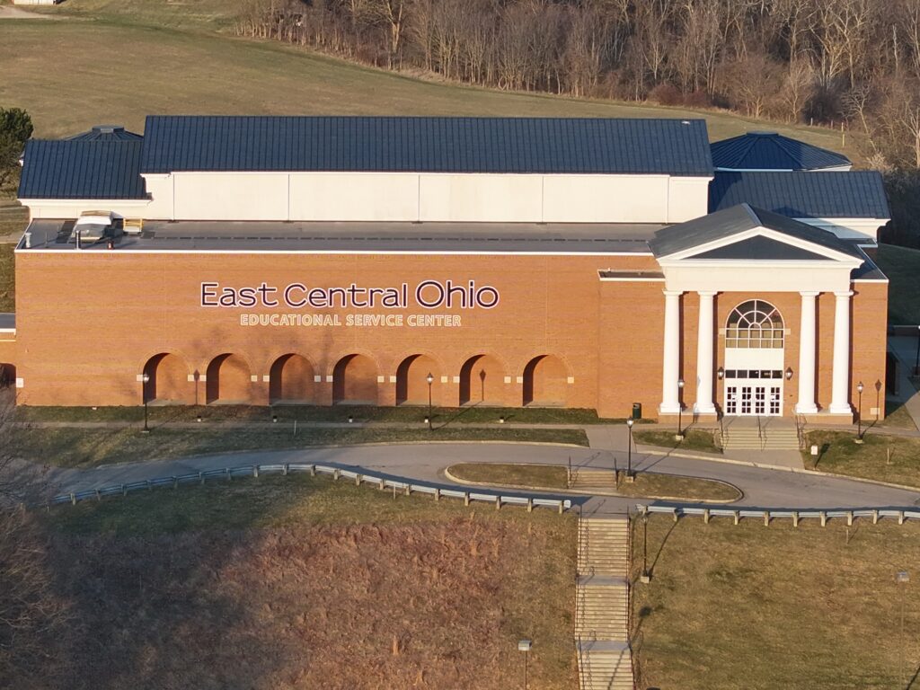 Aerial exterior view of the East Central Ohio Educational Service Center, a large brick building with a navy metal roof, decorative arched facade, white columned entrance, and circular drive, surrounded by green grounds and wooded hills.