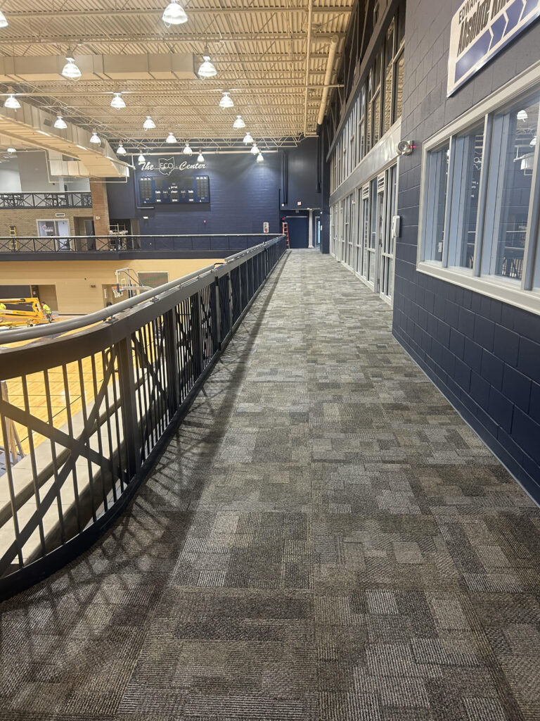 Looking down the newly carpeted upper-level walking track at The ECO Center, with patterned carpet tiles, a metal railing, navy-painted walls, and the gymnasium floor visible below.
