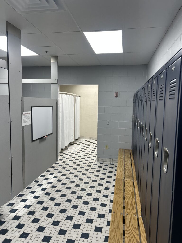 Renovated locker room hallway at The ECO Center featuring a row of new gray metal lockers, wooden benches, white curtained shower stalls, and a classic black-and-white checkered tile floor.