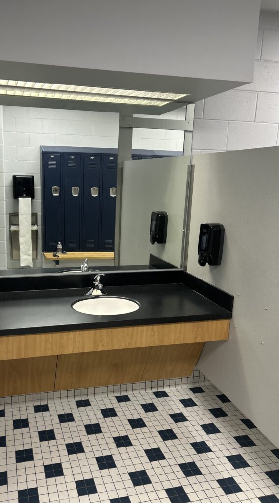 Renovated locker room vanity area at The ECO Center featuring a dark countertop sink, large mirror, navy lockers, and classic black-and-white checkered tile flooring.