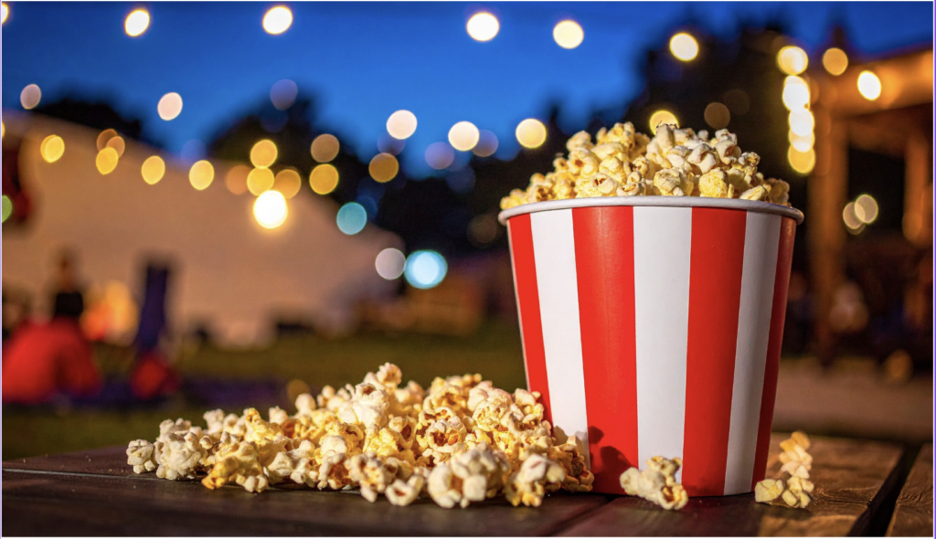 A red and white striped popcorn bucket overflowing onto a wooden table at an outdoor evening event, with string lights bokeh in the background.