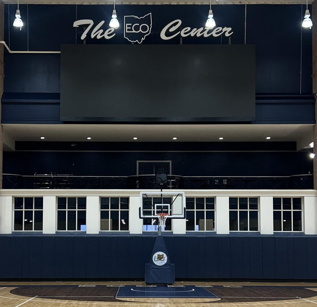 View from the gymnasium floor looking toward The ECO Center's renovated upper bowl, featuring navy wall padding, a large display screen, basketball hoop, and the facility's signature logo mounted above center court.
