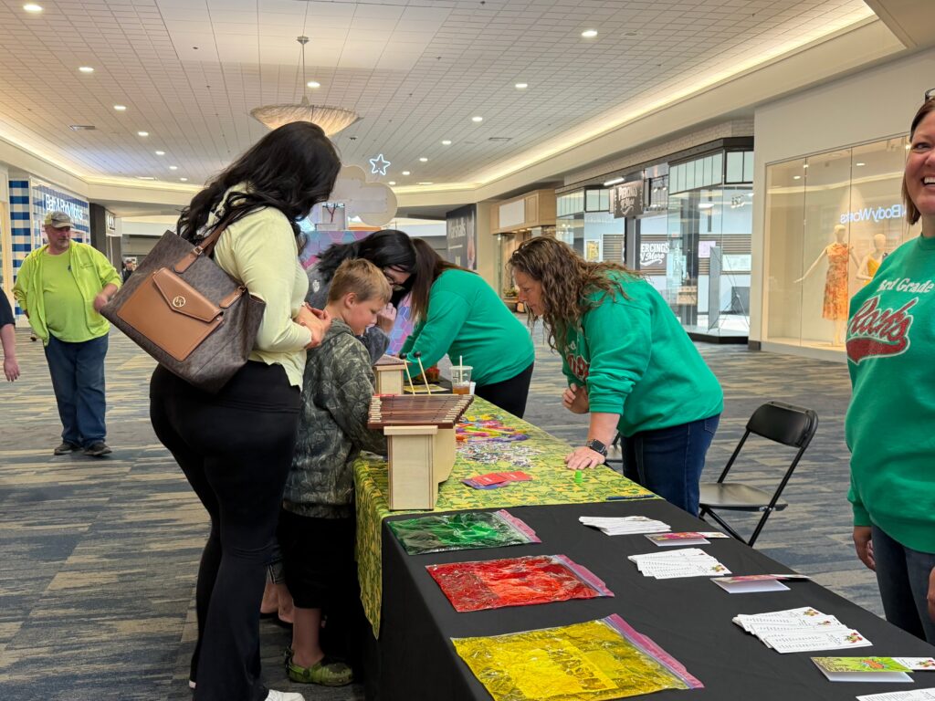 Families browse a colorful activity table staffed by volunteers in green shirts at an indoor reading festival.