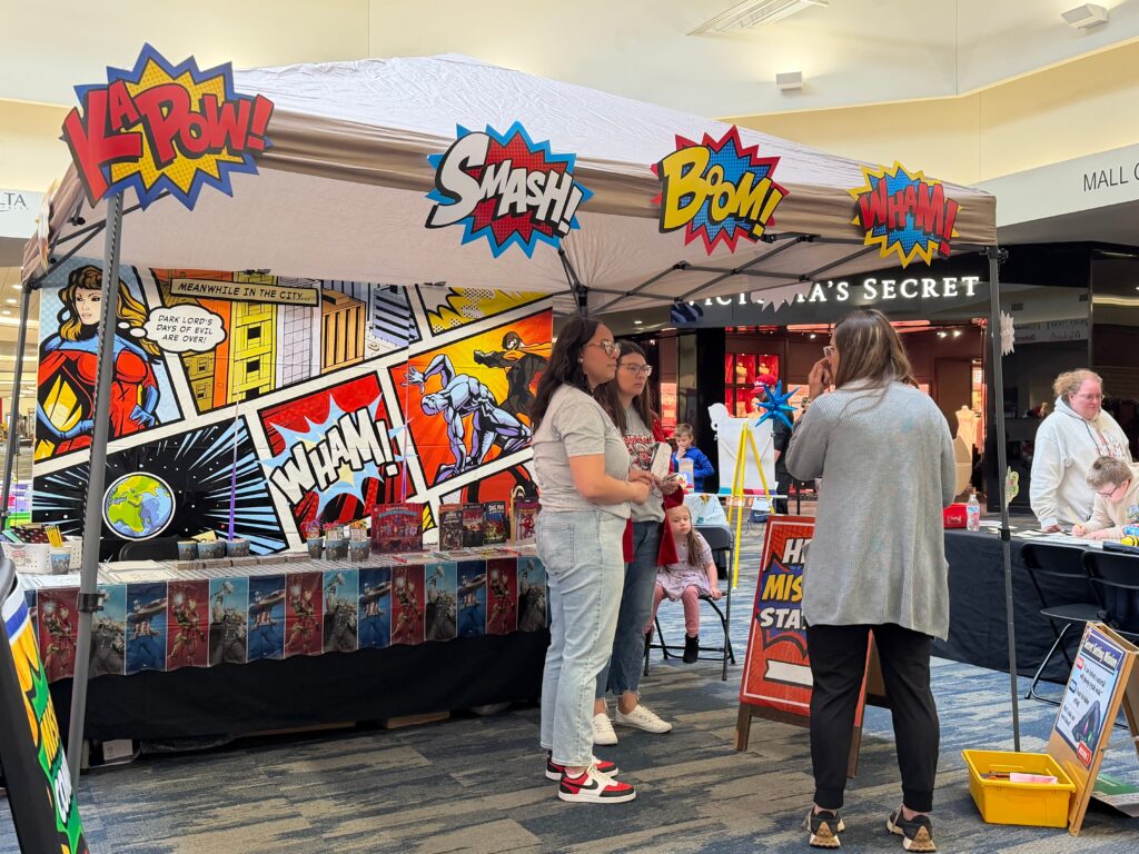 Families visit a superhero-themed booth decorated with comic book style signs including "Pow," "Smash," and "Boom."