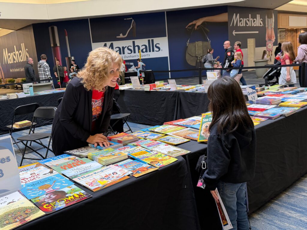A woman assists two children at a table filled with colorful picture books at an indoor festival booth.