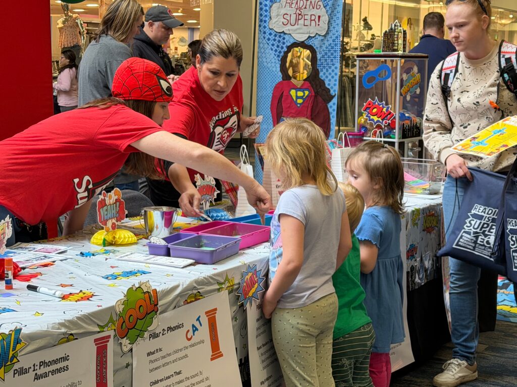 Children and adults gather around a bright, colorful activity table participating in a hands-on literacy activity.
