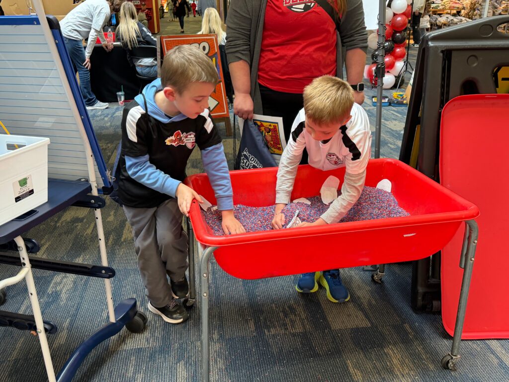 Two young boys dig through a red sensory bin filled with small objects as part of a reading activity.