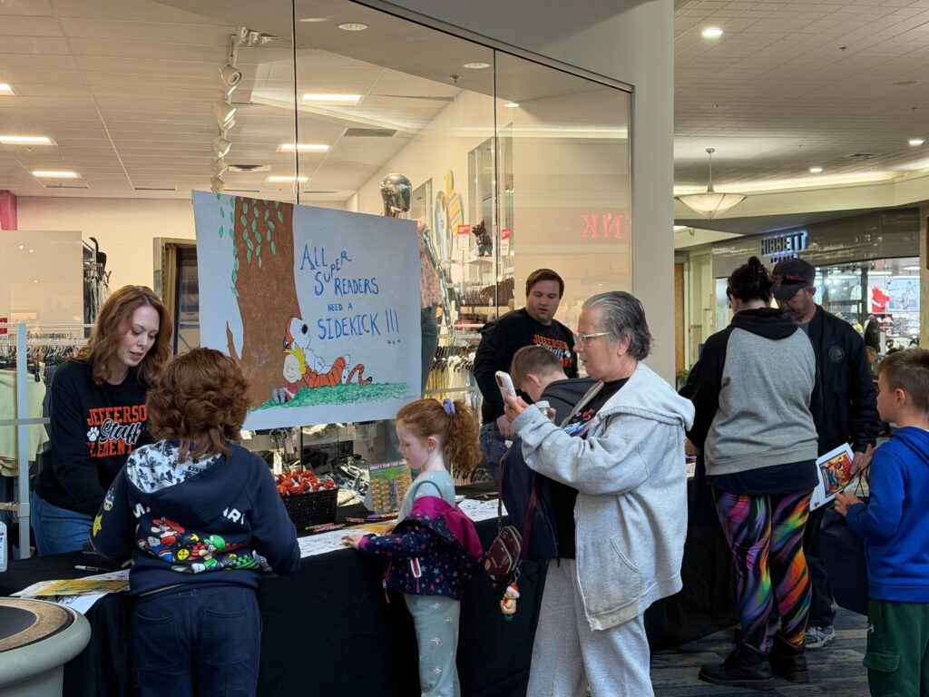 Families interact with volunteers at a booth featuring handmade signage reading "All Students Can Read at Scratch It."