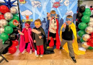 Three young children dressed in superhero capes and masks pose in front of a colorful balloon and photo backdrop.